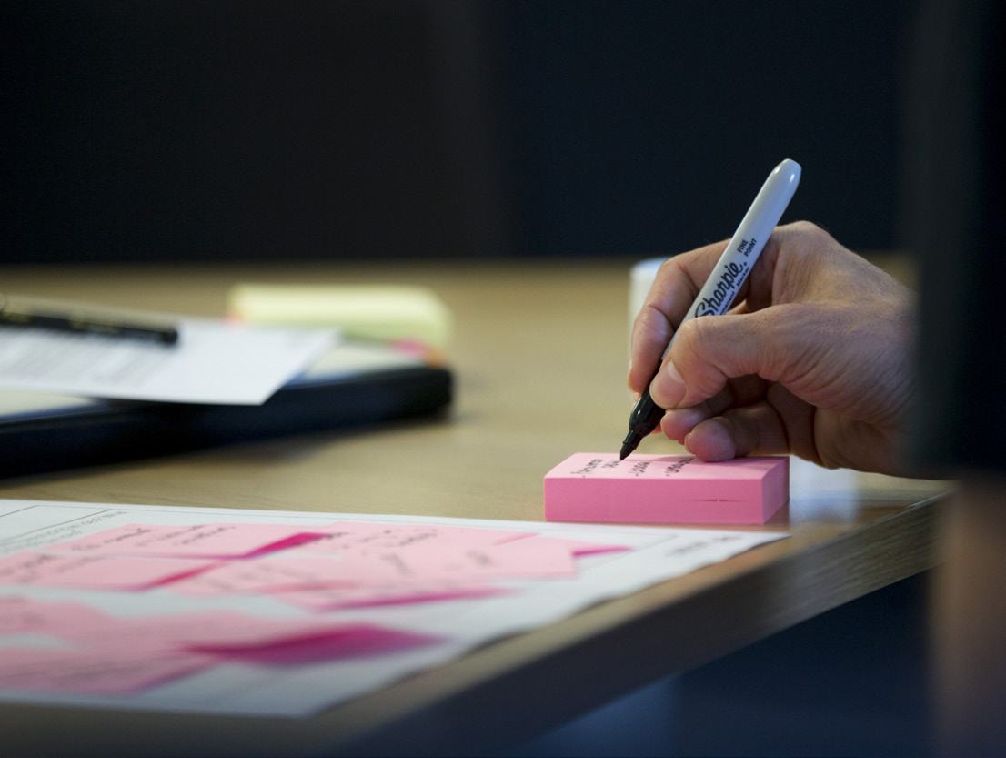 Close-up of a hand writing with a black Sharpie on a stack of pink sticky notes, suggesting a brainstorming or note-taking activity.