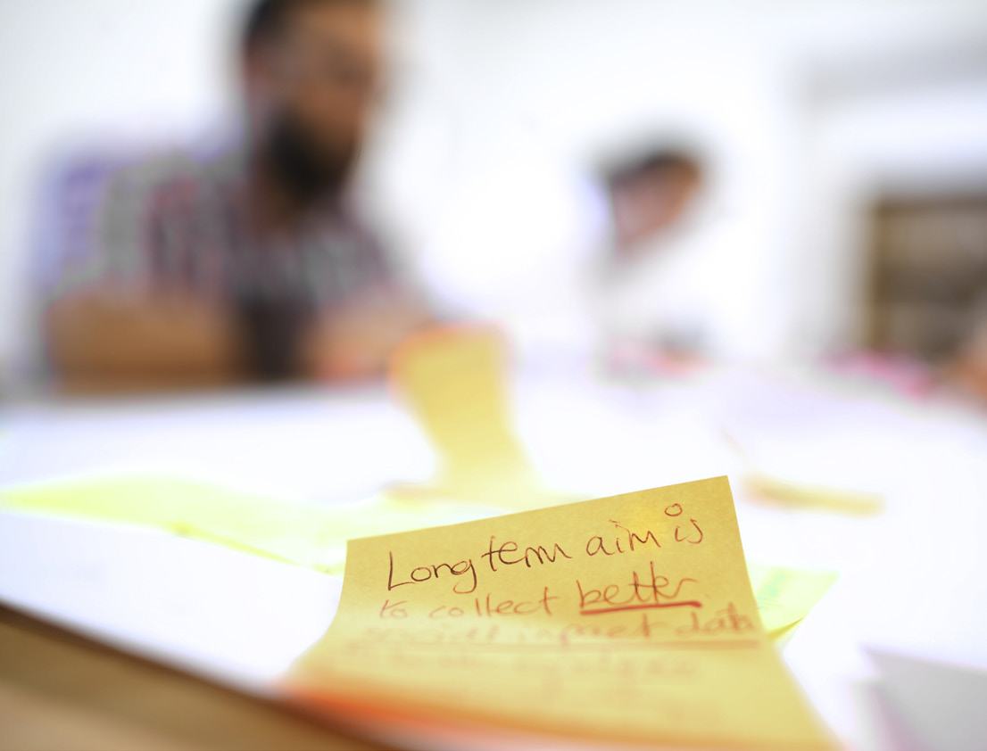 Close-up of a yellow sticky note with handwritten text: "Long term aim is to collect better data."