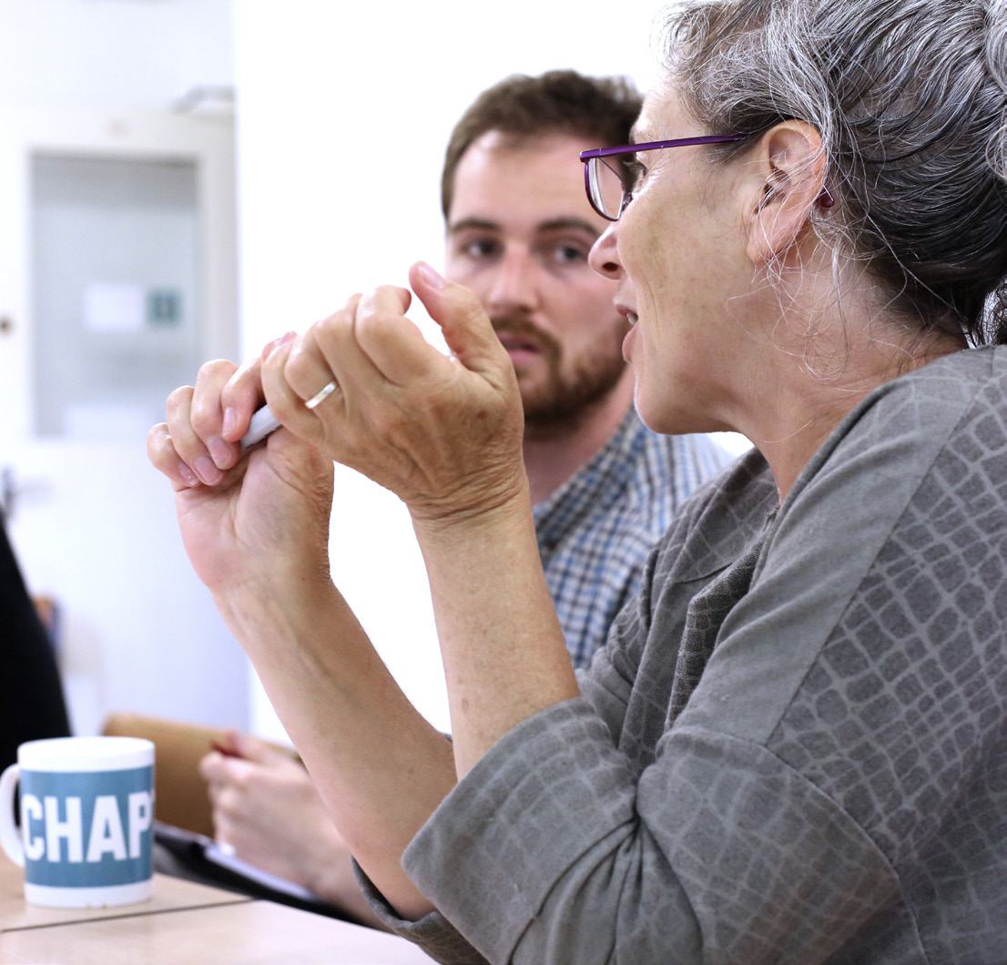 A woman gesturing while speaking passionately in a meeting, with a colleague listening in the background.