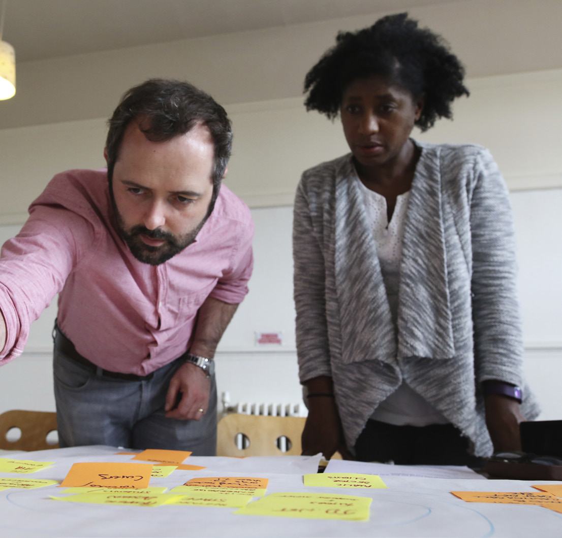 A man and woman collaborate, leaning over a table covered with a large sheet of paper and numerous yellow and orange sticky notes.