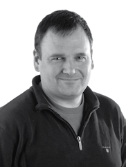 Black and white headshot of a man with short hair, wearing a zipped-up sweater, looking forward and slightly smiling.