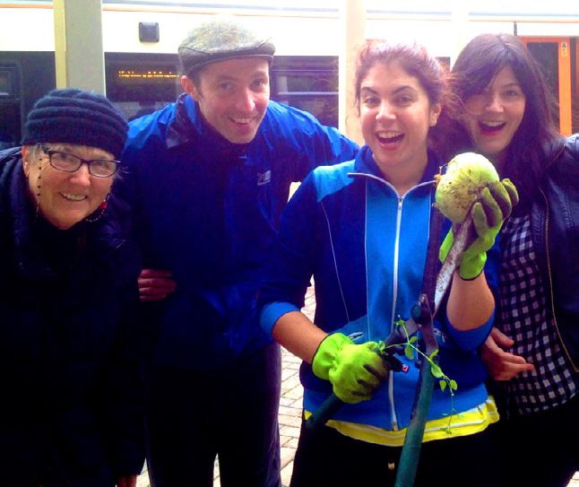 Four smiling people, wearing casual clothes, hold a large green gourd and gardening tools.