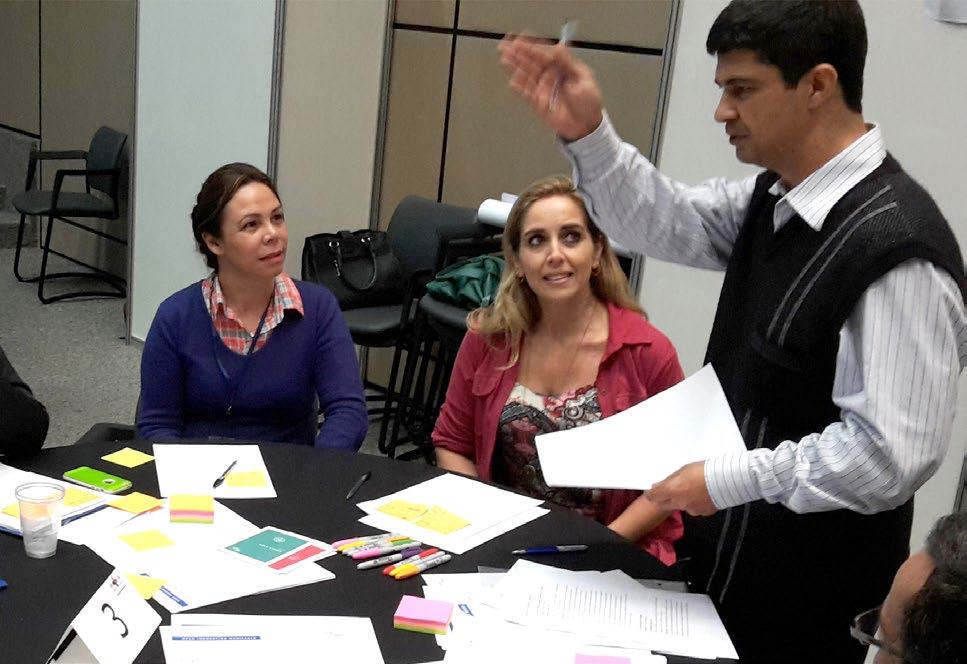 A man presents or instructs two women seated at a table, holding papers and gesturing, in a meeting or workshop setting.