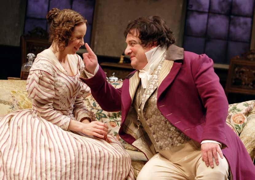 Two actors in period costumes on a sofa; a woman in a striped dress looks at a man in a purple coat who gestures and smiles at her.