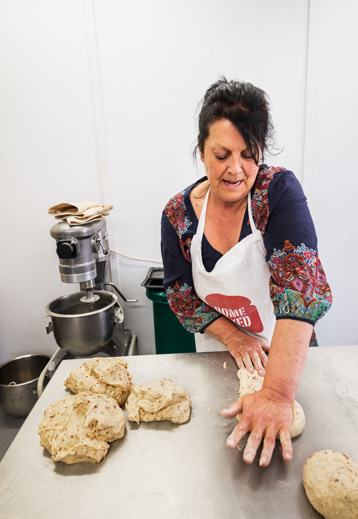 A woman in an apron kneading bread dough on a metal table, with a large stand mixer nearby.