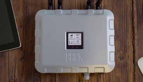 Top-down view of a light gray BRCK device with a small screen showing battery, signal, and user count indicators, placed on a wooden table.