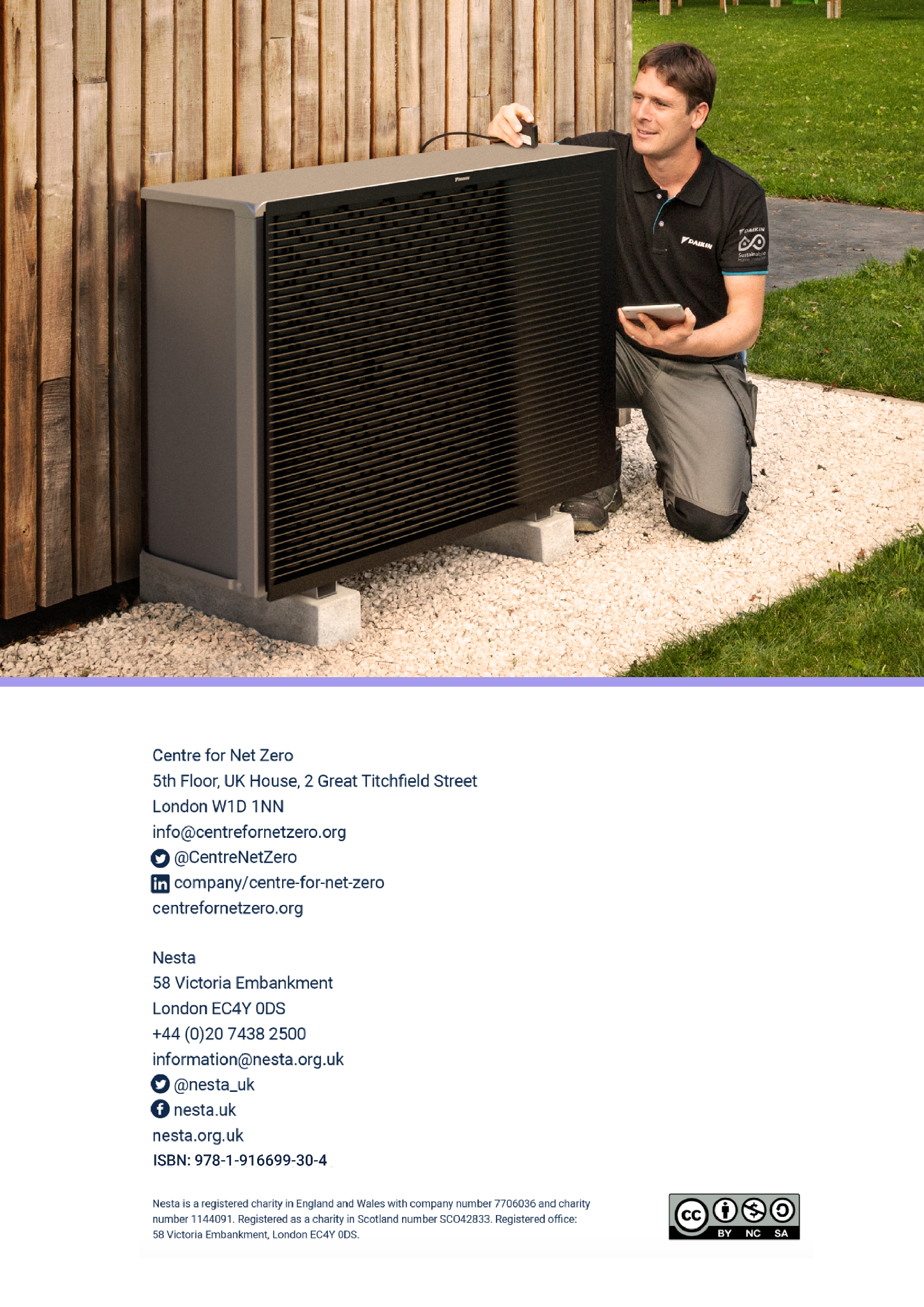 A technician kneels beside a large outdoor heat pump unit, looking at it while holding a tablet.