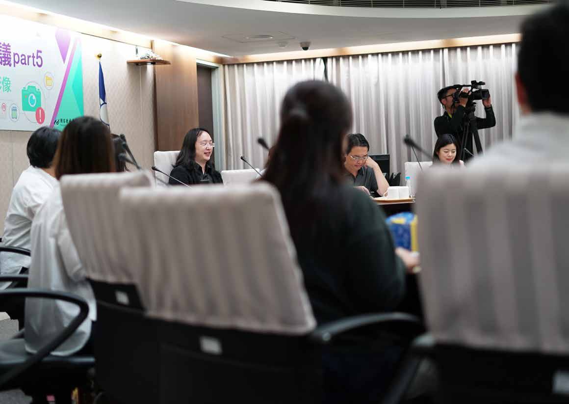 People attending an indoor meeting or presentation, with a speaker at the head of the table and a cameraman filming in the background.
