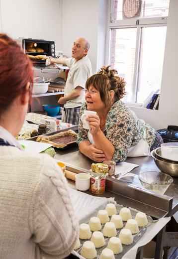 People working in a bustling bakery kitchen, including a man at an oven and a woman with a mug at a table of pastries.
