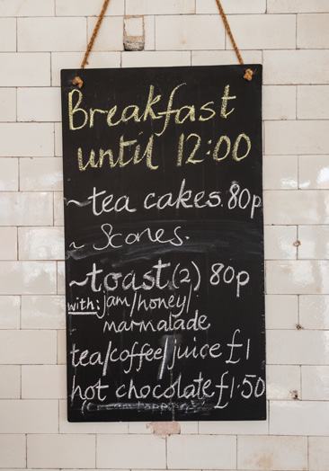A chalk board menu listing breakfast items and prices, including tea cakes, scones, toast, and drinks.