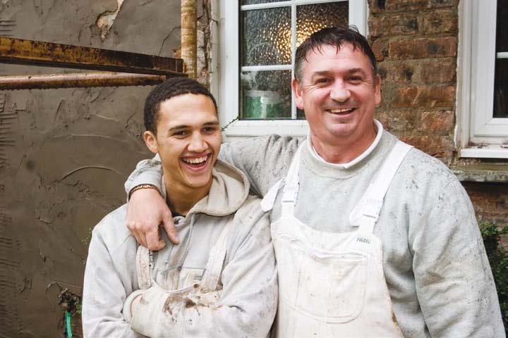 Two smiling men, covered in plaster or paint, stand arm-in-arm in front of a building under renovation.