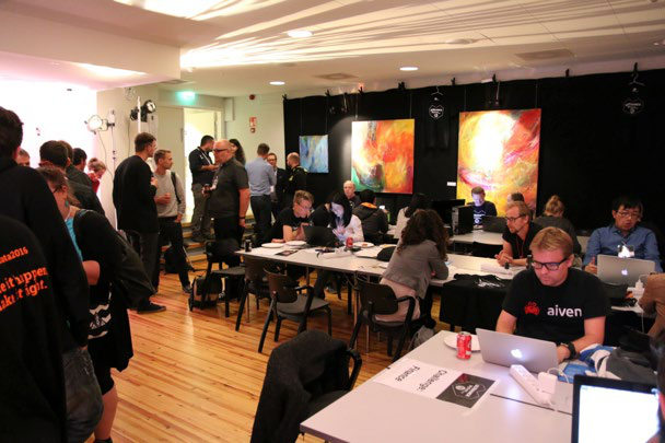 People at a data workshop or hackathon, some standing, some working on laptops at tables, in a brightly lit room with abstract art.