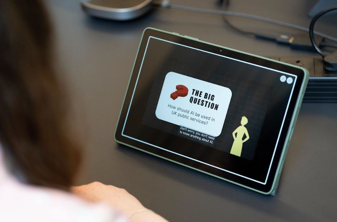 Close-up of a tablet screen displaying the question "THE BIG QUESTION: How should AI be used in UK public services?". Text also says "Don't worry, you don't need to know anything about AI." A person's shoulder is visible in the foreground.