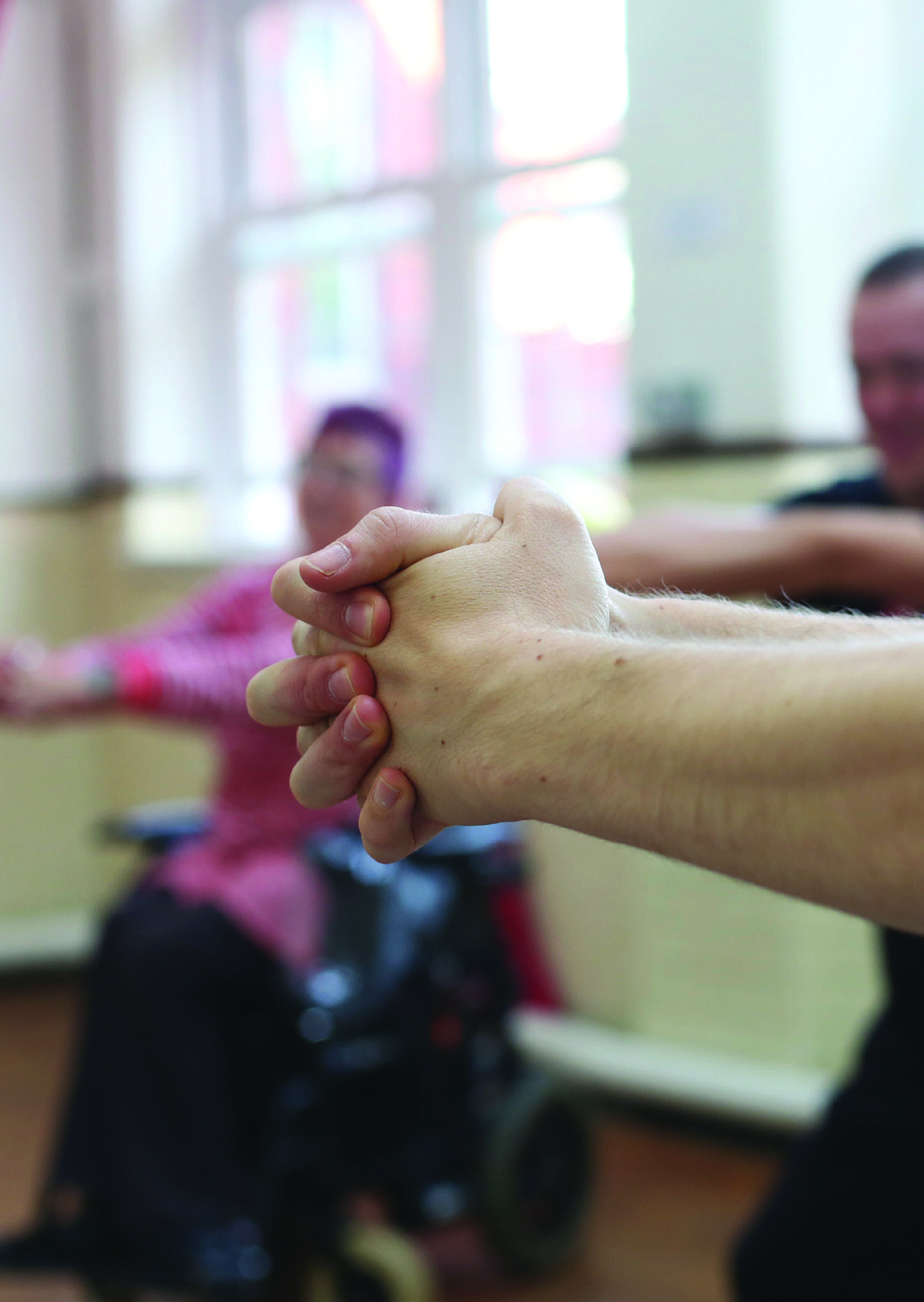 Close-up of clasped hands during an exercise, with people including one in a wheelchair in the blurry background.