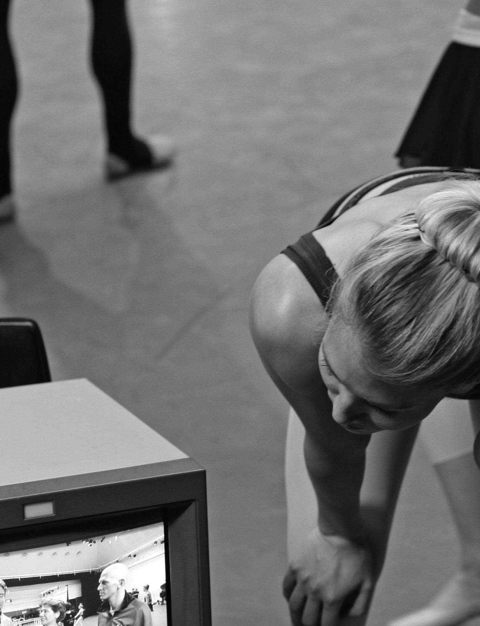 Dancer stretching with a monitor displaying a performance or coaching session in the foreground.
