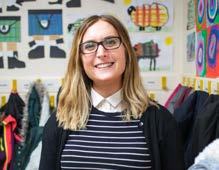 Smiling woman with glasses in a classroom setting, with colorful children's artwork displayed on the wall behind her.