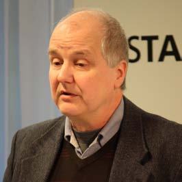 Close-up portrait of an older man with short grey hair, wearing a jacket and sweater, speaking at an event with Nesta branding.