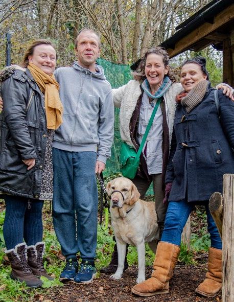 Four smiling people and a yellow Labrador dog pose outdoors in a wooded area, dressed in warm clothing.