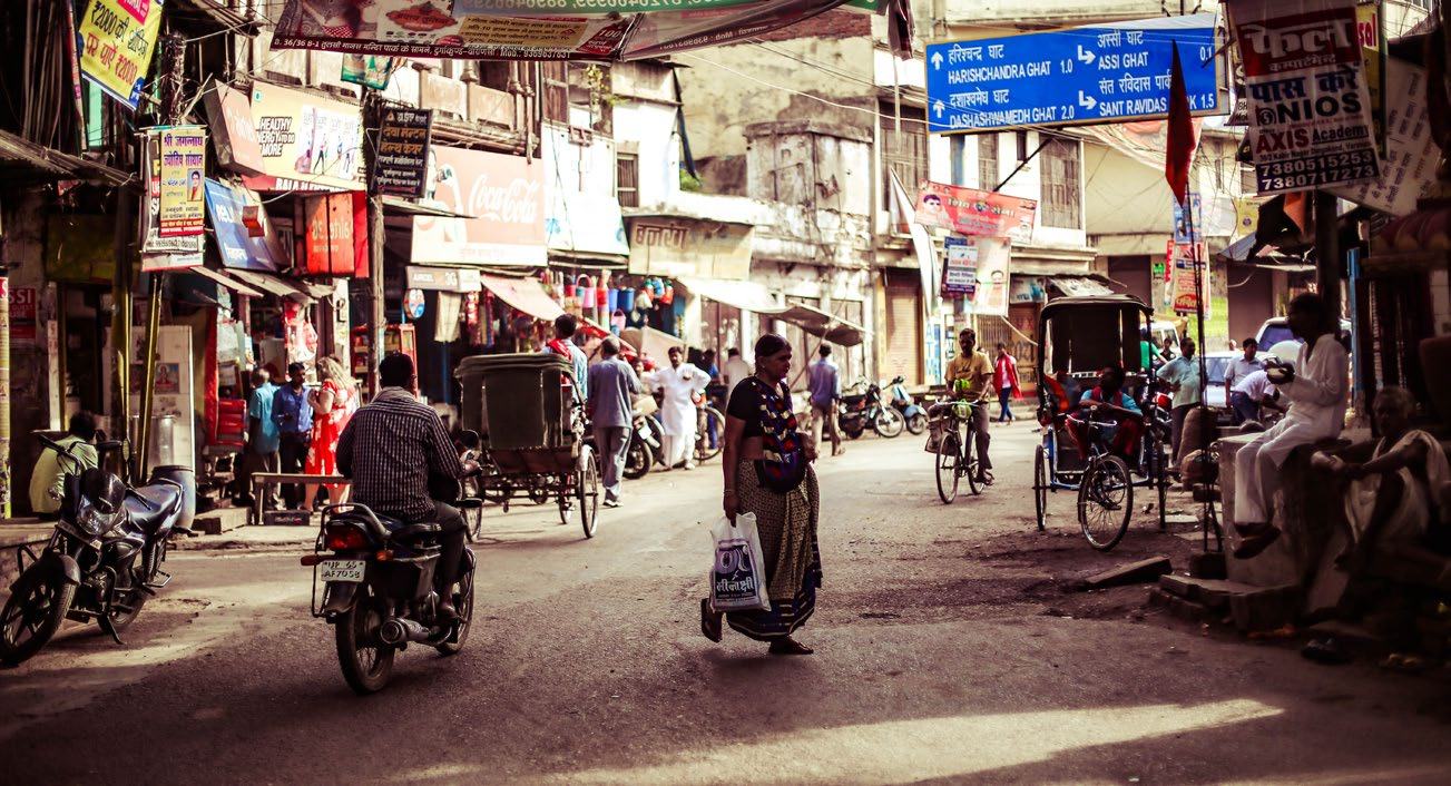 A bustling street scene in an Indian town with shops, motorcycles, auto-rickshaws, pedestrians, and street signs.