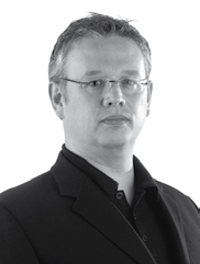 Black and white headshot of a man with short hair and glasses, wearing a collared shirt and jacket, looking forward.