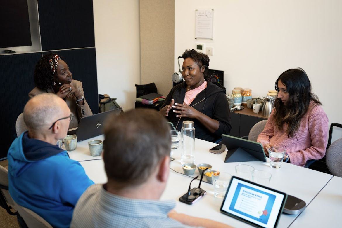 Five people engaged in a discussion around a table, with tablets and laptops, one tablet displaying 'Introducing Risks' in a deliberation session.