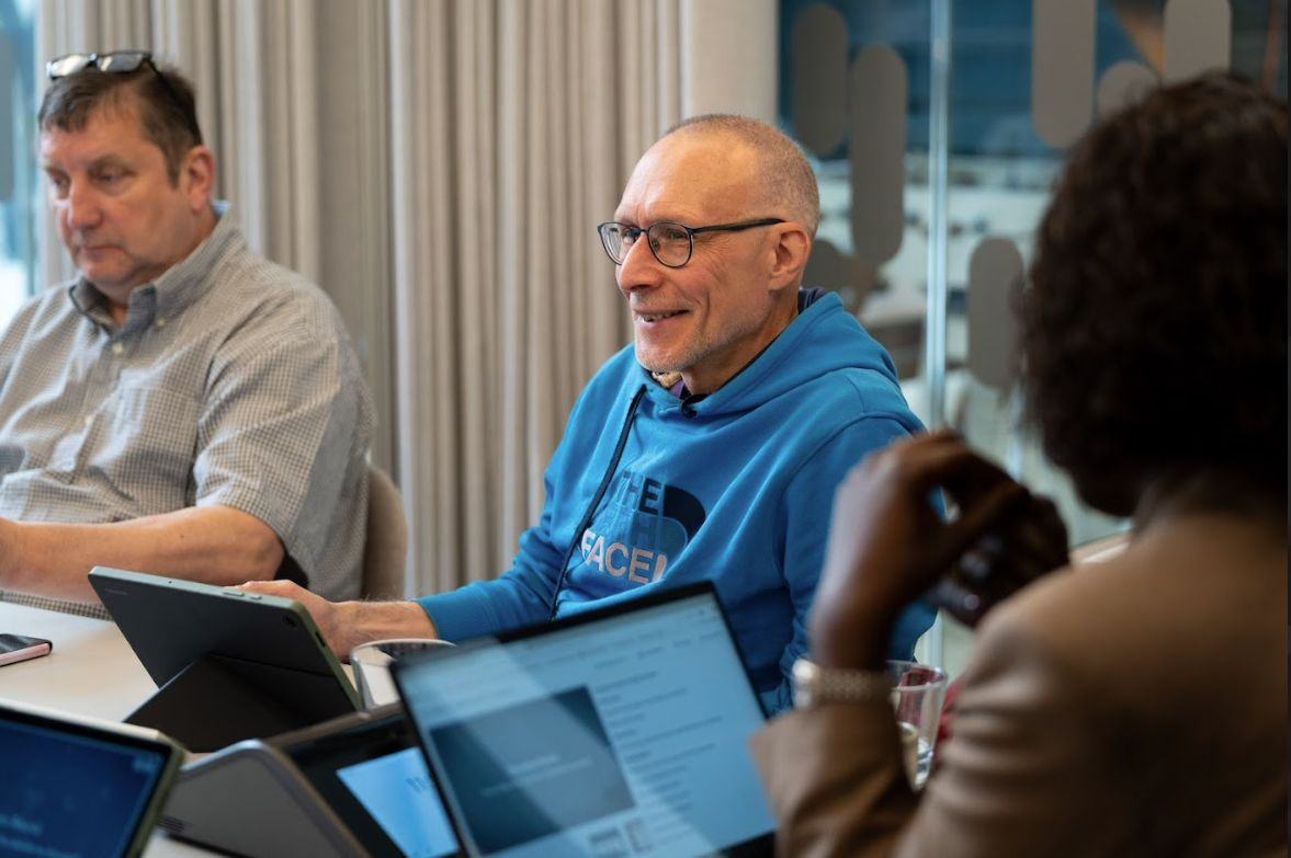 A smiling man, a participant in a deliberation session, with laptops and tablets visible on the table in front of him.