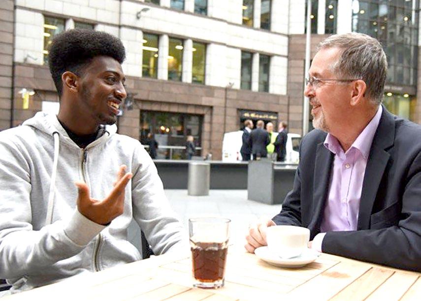 Two men having a conversation at an outdoor cafe table, potentially representing mentorship or a business meeting.