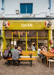 Exterior of Okan Japanese restaurant with people dining at outdoor picnic tables under yellow signage.