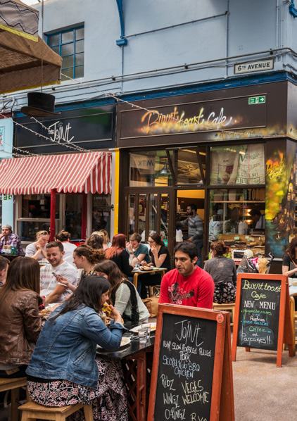 People dining outdoors at cafes in a busy market, with visible menu boards showing items like pulled pork and ribs.