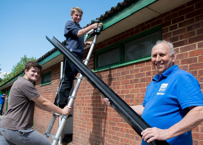 Three males, including an older mentor, installing a gutter on a brick building while smiling and working together.