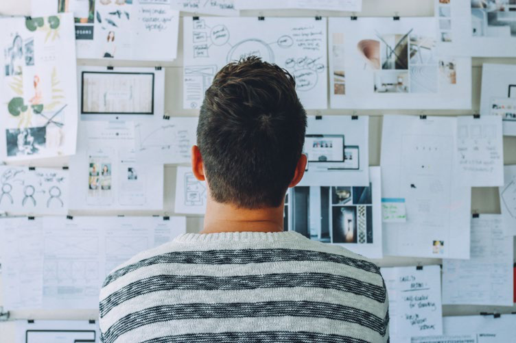 Person seen from behind, looking at a wall covered with numerous papers, notes, and images, suggesting brainstorming or project planning.