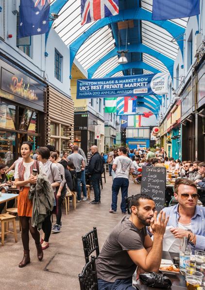 Bustling covered market street with cafes, shops, and people, featuring a banner proclaiming 'Brixton Village & Market Row UK's Best Private Market'.