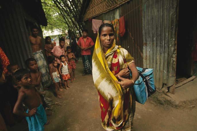 A woman in traditional attire, carrying a blue bag, walks past children and corrugated metal homes in an informal settlement.