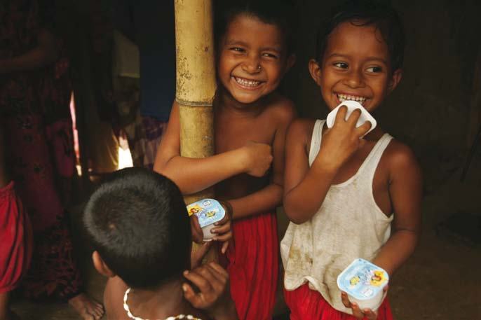 Three young children happily smiling and eating small cups of yogurt or ice cream in an indoor setting.