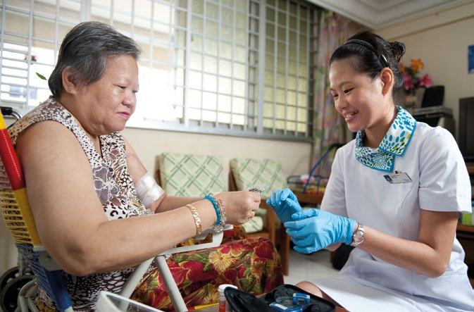 A smiling nurse in blue gloves assists an elderly woman in a wheelchair, demonstrating healthcare or elder care interaction.