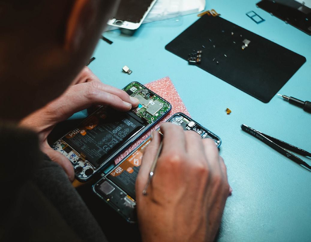 Close-up of hands repairing a smartphone, with internal components visible on a blue workbench alongside tools.