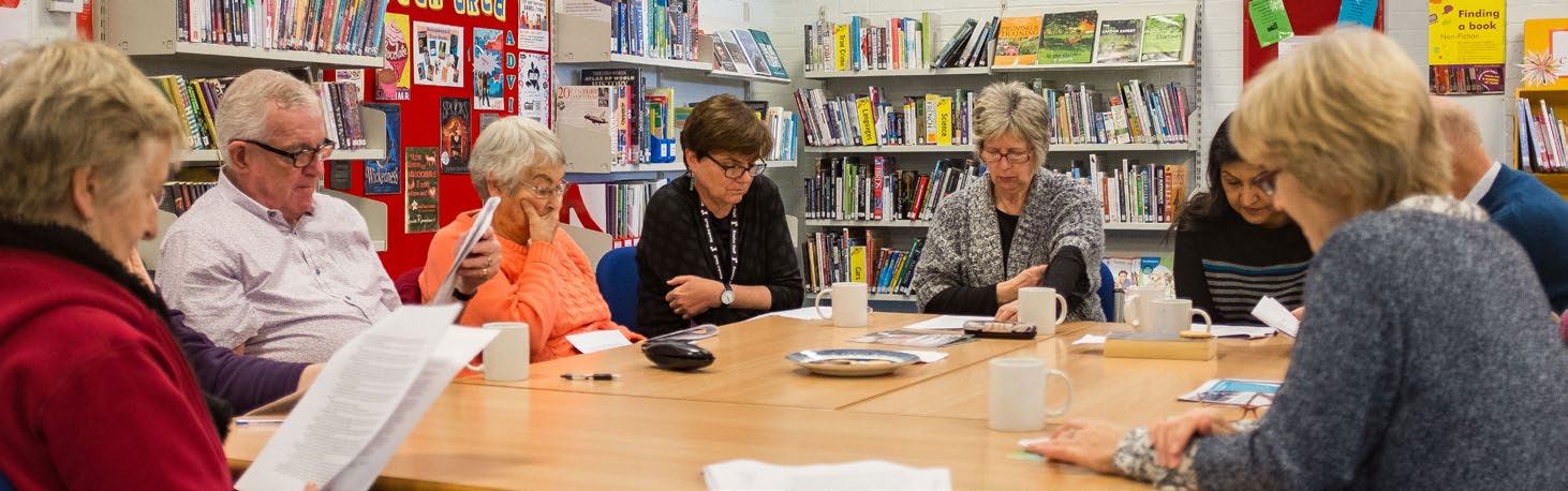 A diverse group of adults sitting around a table in a library, reading documents and engaging in a discussion with bookshelves in the background.