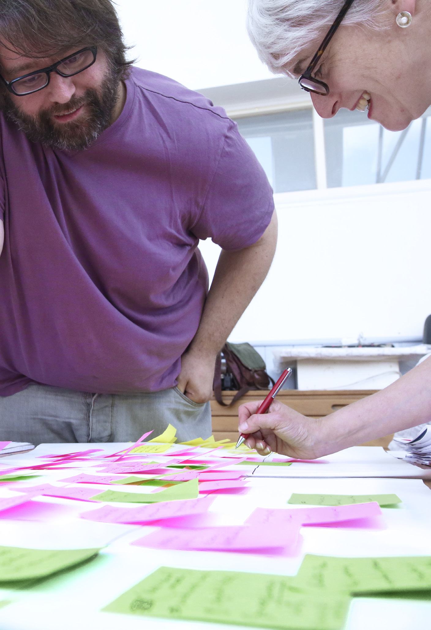 Two people, a man and a woman, actively collaborating and writing on numerous pink and green sticky notes on a table.