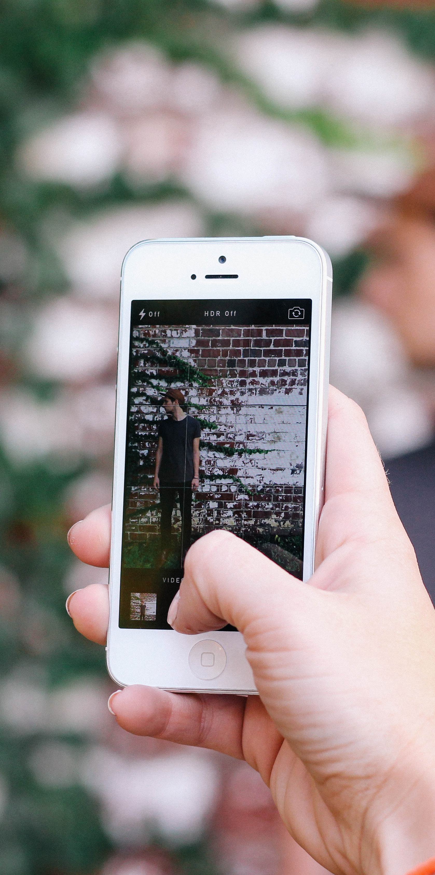 Hand holding a smartphone displaying the camera app interface, showing settings like flash and HDR, while framing a person against a brick wall.