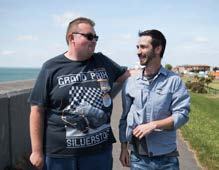 Two men, one wearing sunglasses, talking and smiling outdoors by a wall with a coastline in the background.