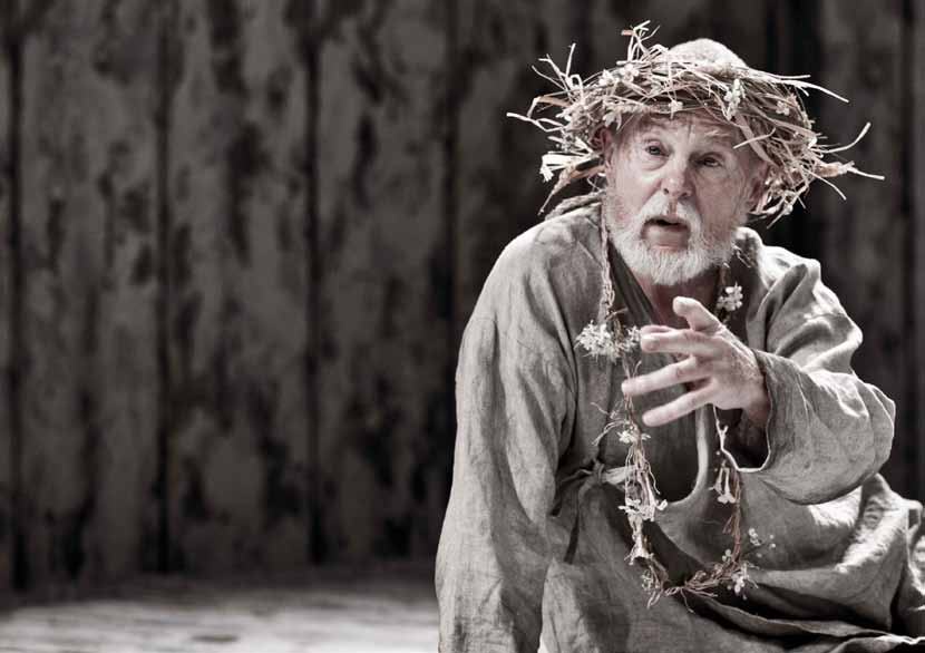 Older man with white beard and twig wreath, wearing a rustic costume, gestures on a stage with a dark wooden background.