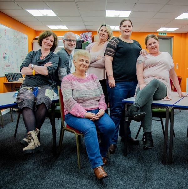 Six people, five women and one man, smile while posing in a classroom with orange walls and tables.