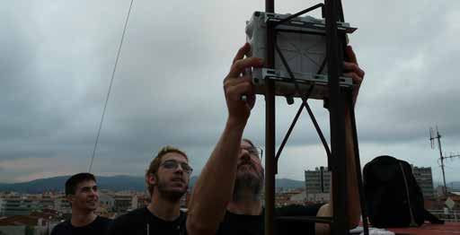 Three individuals on a rooftop installing a small, cube-shaped sensor or satellite device onto a metal pole against a cloudy sky.