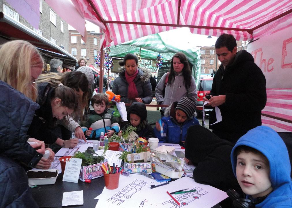 Children and adults participate in a craft activity, drawing and coloring under a striped market tent.