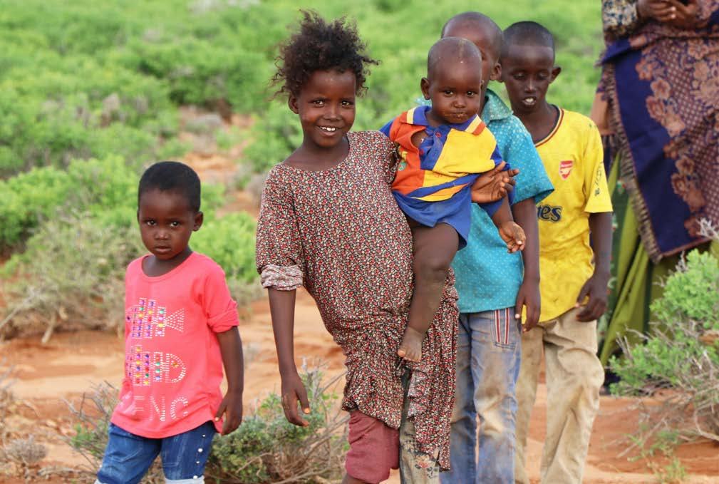 A group of children, some barefoot, smiling and standing in a semi-arid landscape with sparse vegetation, depicting community in a rural setting.