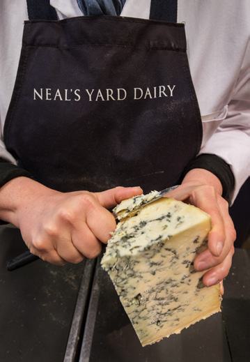 Close-up of hands in a "Neal's Yard Dairy" apron, slicing a wedge of blue cheese.