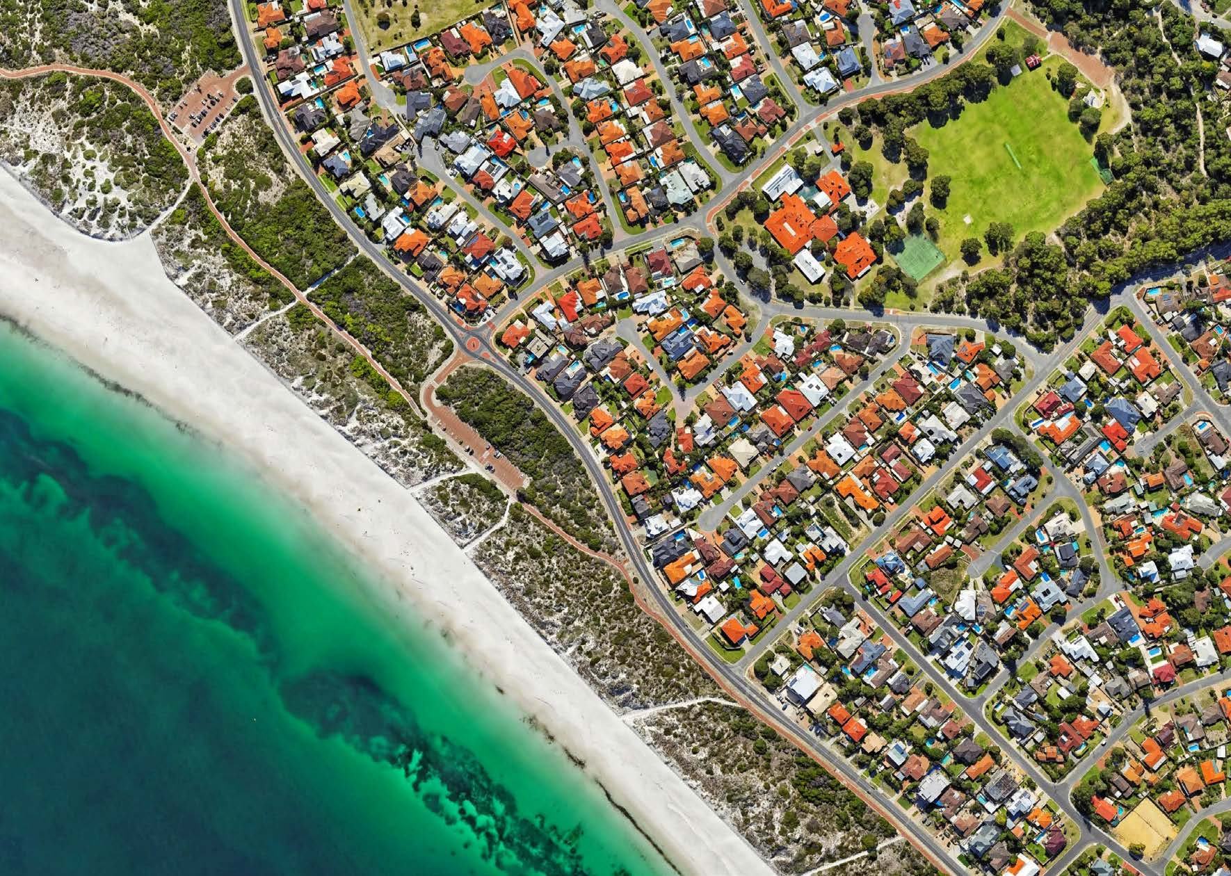 Aerial view of a coastal residential area with houses, roads, a green oval, a white sand beach, and turquoise ocean.
