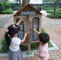 Two young children reach into a small public book-sharing box shaped like a house in a park, engaging with community literacy.
