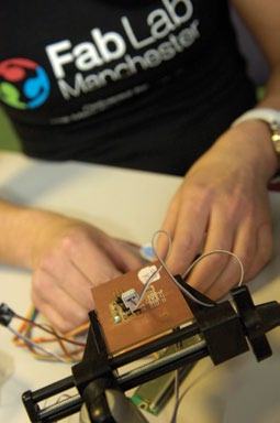 A person’s hands are shown soldering wires to a small circuit board, with a 'Fab Lab Manchester' t-shirt visible.