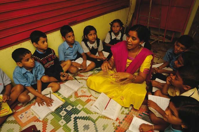A woman in traditional dress teaching a group of young children sitting on the floor around her, with open books.